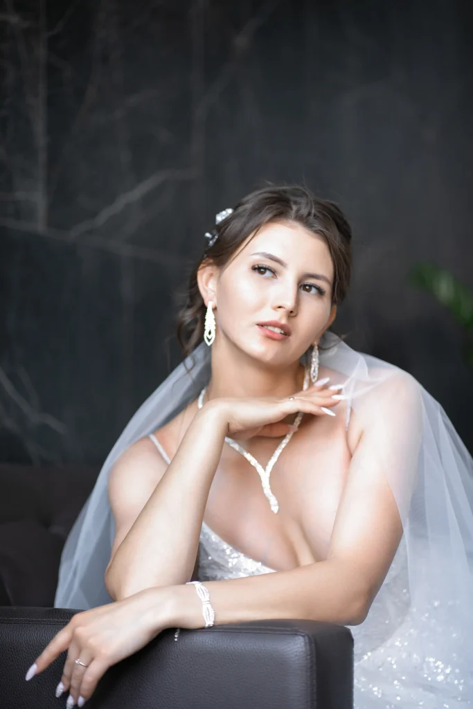 viktoriya i ildar . svadebnaya seriya . velkosta 0112 Невеста в свадебном платье, сидящая на диване перед стеной. На ней белое платье с фатой и тиарой, а ее волосы уложены в элегантную прическу. Ее макияж едва заметен, а выражение лица выражает радость и предвкушение.